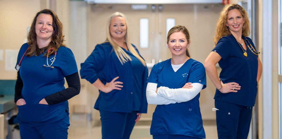 four nurses in blue scrubs standing in hospital hallway with crossed arms in a power pose