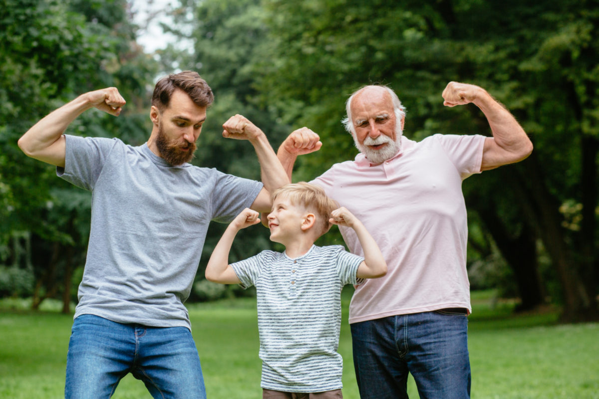 Portrait of happy family - grandpa, father and his son smiling and showing their muscles outdoor in park on background. Three different generation concept.